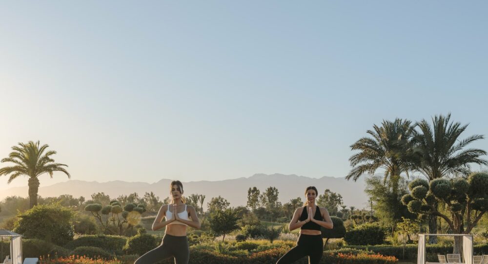 Deux femmes en posture de yoga au lever du jour au Fairmont Royal Palm Marrakech, jardins et Atlas en arrière-plan