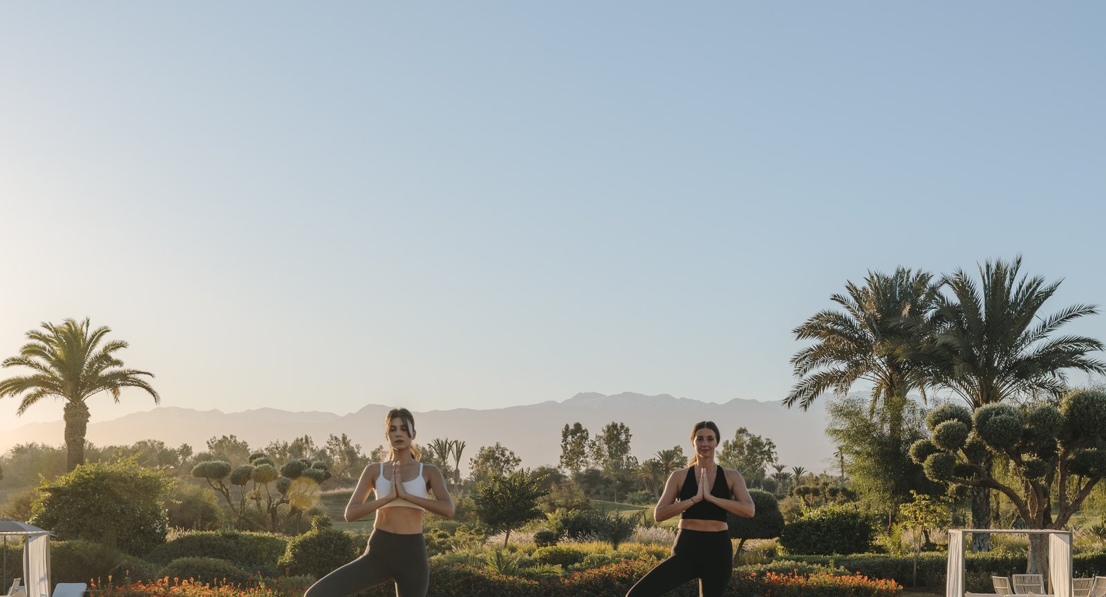 Deux femmes en posture de yoga au lever du jour au Fairmont Royal Palm Marrakech, jardins et Atlas en arrière-plan